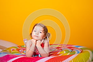 Happy little girl in swimming suit lying on colorful inflatable mattress lollipop on yellow background.