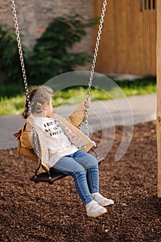 Happy little girl swing on a swing in the specially designated place in the park. Softner from sawdust underfoot