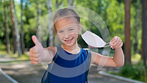 Happy little girl takes off the mask and shows thumb outdoors. Joyful smiling schoolgirl with a backpack pushes the mask photo