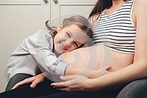 Happy little girl touching her pregnant mom tummy. Pregnant mother and daughter spending time together at home. Sweet daughter