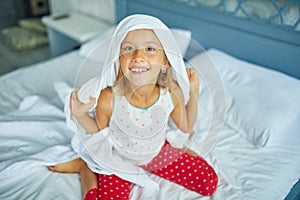 Happy little girl wakes up, joyful, blanket on the head, from sleep on a big and cozy bed white linen in the at home