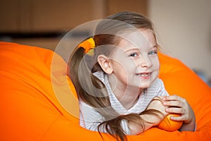 Happy little girl wallows in an orange bean bag chair with a ripe orange fruit