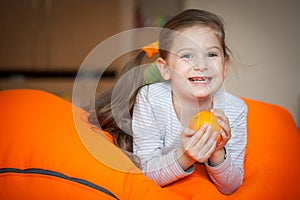 Happy little girl wallows in an orange bean bag chair with a ripe orange fruit