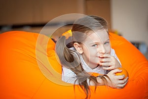 Happy little girl wallows in an orange bean bag chair with a ripe orange fruit