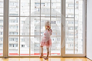 Happy little girl wearing pink dress eating lollipop while standing in front of big window