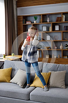 Happy little girl wearing red earphones dancing and singing using smartphone like microphone. Cute child having fun