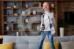 Happy little girl wearing red earphones dancing and singing using smartphone like microphone. Cute child having fun