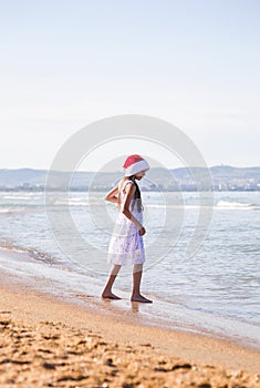 Happy little girl wearing a red Santa hat walking through the sea water on clear sunny day, vertical