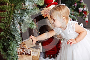 Happy little girl in white dress puts firewood in the Christmas wooden brown decorated room interior