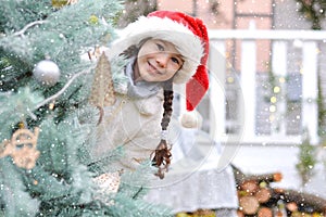 A happy little girl in a white fur coat and santa hat peeks out from behind a Christmas tree against the backdrop of a New Year`s