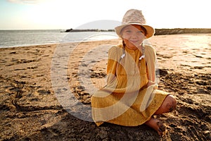 Happy little girl in yellow cute dress and summer hat enjoying sunny day at the beach, sitting on the sand and smiling to the