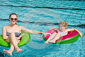 Happy little girl and young mother playing with colorful inflatable rings in outdoor swimming pool on sunny summer day