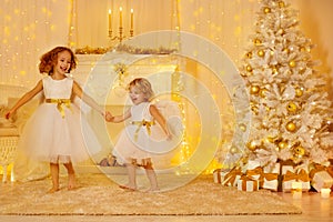 Happy Little Girls Playing Next to Christmas Tree with Gifts in White Home Room Interior with Fireplace Candles and Golden Xmas