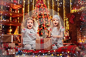 Happy little girls wearing Christmas pajamas open gift box by a fireplace in a cozy dark living room on Christmas eve.