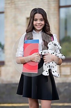 Happy little kid back to school in uniform holding books and toy dog in schoolyard, knowledge day