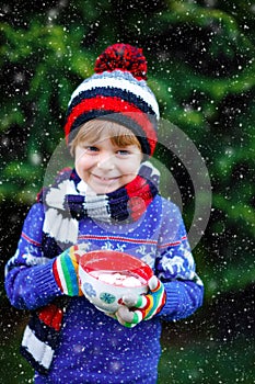 Happy little kid boy drinking hot cocoa and chocolate drink and marshmallows. Funny child in winter sweater, cap, long