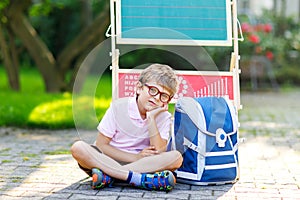 Happy little kid boy with glasses sitting by desk and backpack or satchel