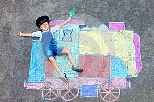 Happy little kid boy having fun with train or steam locomotive picture drawing with colorful chalks on ground. Children