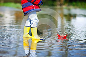 Happy little kid boy in yellow rain boots playing with paper ship boat by huge puddle on spring or autumn day