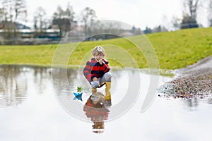 Happy little kid boy in yellow rain boots playing with paper ship boat by huge puddle on spring or autumn day