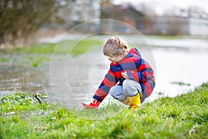 Happy little kid boy in yellow rain boots playing with paper ship boat by huge puddle on spring or autumn day. Active