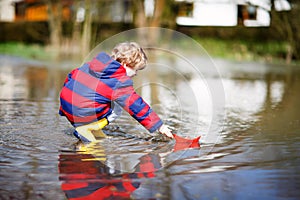 Happy little kid boy in yellow rain boots playing with paper ship boat by huge puddle on spring or autumn day. Active