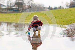 Happy little kid boy in yellow rain boots playing with paper ship boat by huge puddle on spring or autumn day. Active