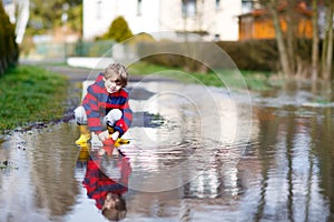 Happy little kid boy in yellow rain boots playing with paper ship boat by huge puddle on spring or autumn day. Active