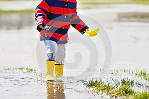 Happy little kid boy in yellow rain boots playing with paper ship boat by huge puddle on spring or autumn day. Active