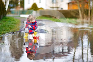 Happy little kid boy in yellow rain boots playing with paper ship boat by huge puddle on spring or autumn day