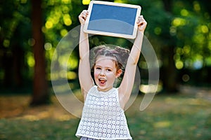 Happy little kid girl holding empty chalk desk in hands. Schoolkid on first day of elementary class. Healthy adorable