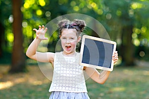 Happy little kid girl holding empty chalk desk in hands. Schoolkid on first day of elementary class. Healthy adorable