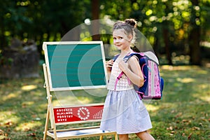 Happy little kid girl standing by desk with backpack or satchel. Schoolkid on first day of elementary class. Back to