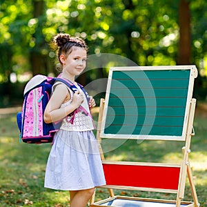 Happy little kid girl standing by desk with backpack or satchel. Schoolkid on first day of elementary class. Back to