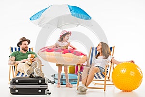 Happy little kid standing with flotation ring between parents sitting on sun loungers