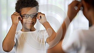 Happy little kid trying on eyeglasses, looking in mirror satisfied good eyesight