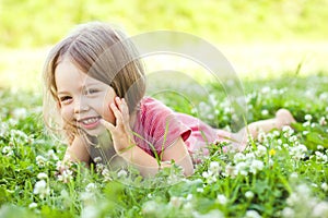 A happy little pretty girl in a light dress is lying on a green grass lawn in the park