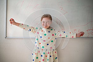 Happy little schoolgirl with down syndrome standing in front of whiteboard and looking at camera