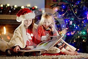 Happy little sisters reading a story book together by a fireplace in a cozy dark living room on Christmas eve