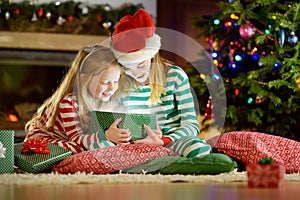 Happy little sisters wearing Christmas pajamas opening gift boxes by a fireplace in a cozy dark living room on Christmas eve.
