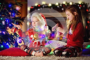 Happy little sisters wearing Christmas pajamas playing by a fireplace in a cozy dark living room on Christmas eve