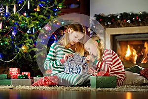 Happy little sisters wearing Christmas pajamas playing by a fireplace in a cozy dark living room on Christmas eve.