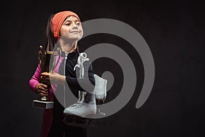 Happy little skater girl champion holds a winner`s cup and ice skates. Isolated on dark textured background.