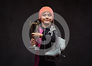 Happy little skater girl champion holds a winner`s cup and ice skates. Isolated on dark textured background.