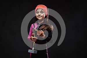 Happy little skater girl champion holds a winner`s cup. Isolated on a dark textured background.