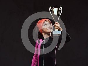 Happy little skater girl champion holds a winner`s cup. Isolated on dark textured background.