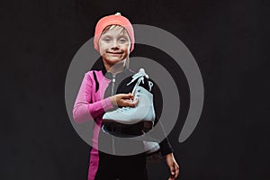 Happy little skater girl dressed in sportswear holds ice skates on a shoulder. Isolated on a dark textured background.