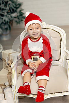 Happy little smiling boy in santa claus costume sits on armchair near christmas tree and holds white candle in hands