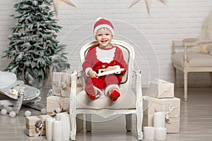 Happy little smiling boy in santa claus costume sits on armchair near christmas tree and holds white candle in hands