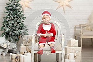 Happy little smiling boy in santa claus costume sitting on armchair near christmas tree and holding christmas gift box